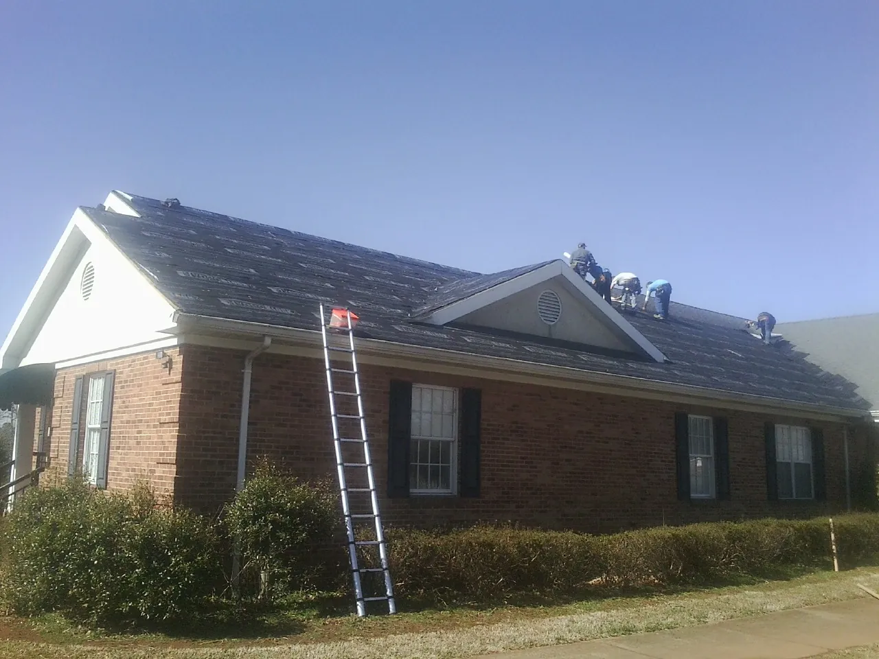 Crew tearing shingles off a brick ranch roof with a ladder set — Emergency Roof Repair work by our crew in Brandt
