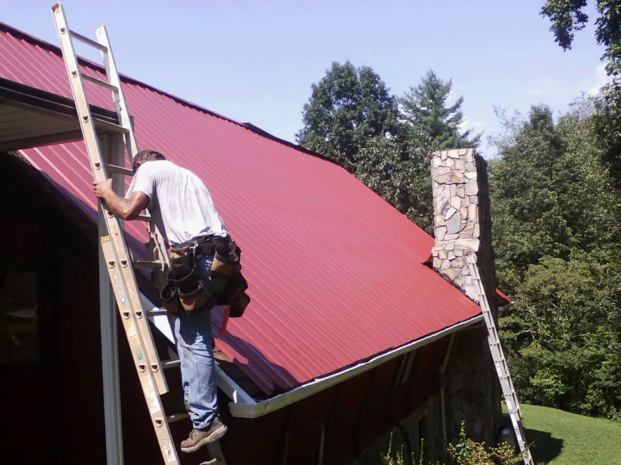 Roofer on a ladder beside a red metal roof and stone chimney — Soffit & Fascia Repair by Roofing Brothers in Brandt
